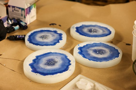 Four freshly poured resin geode coasters in white molds with bright blue and white alcohol ink centers and silver glitter details, sitting on a brown paper-covered table.